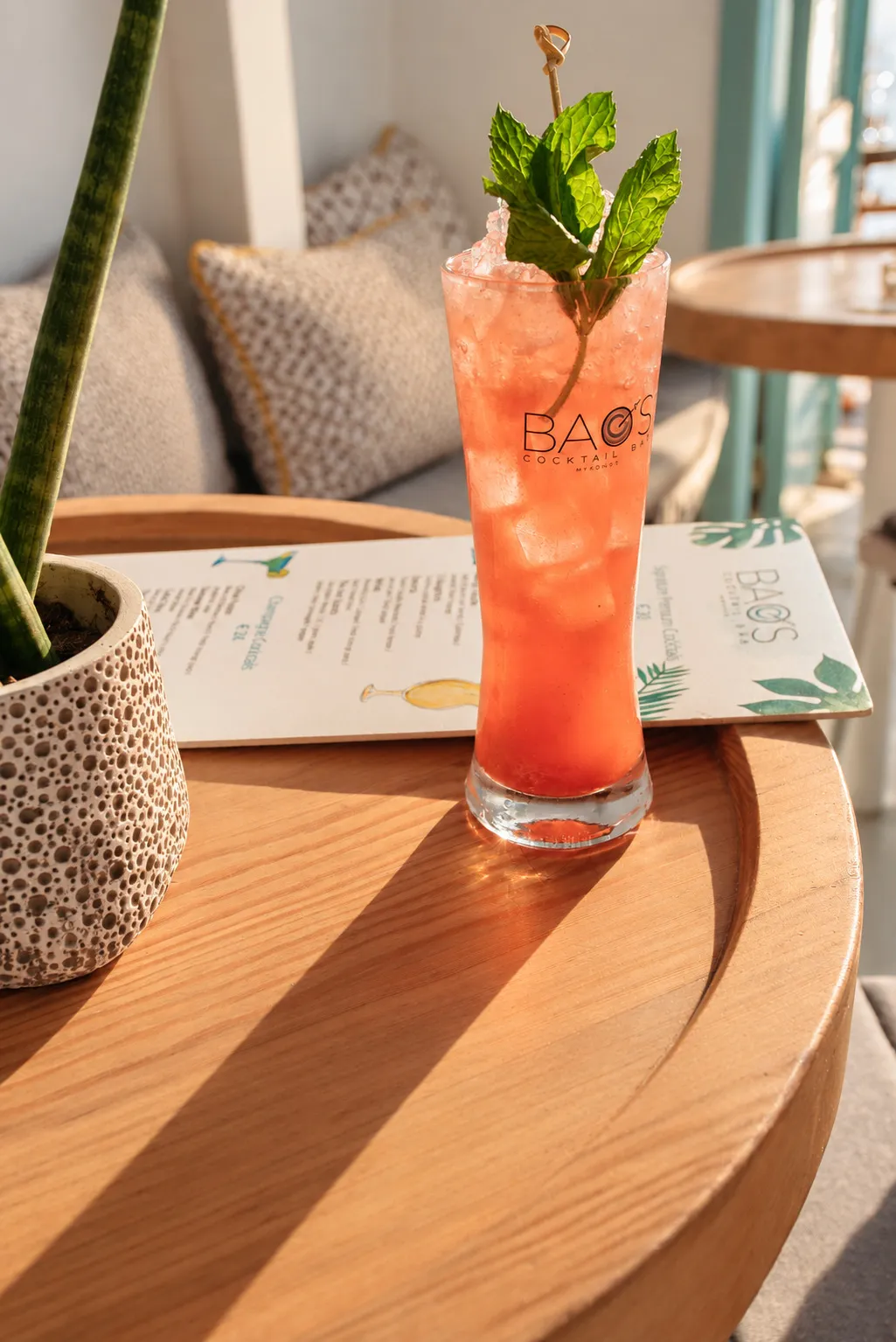 A tall BAO'S signature cocktail on the round wooden table, the printed menu beside it in low sunset light