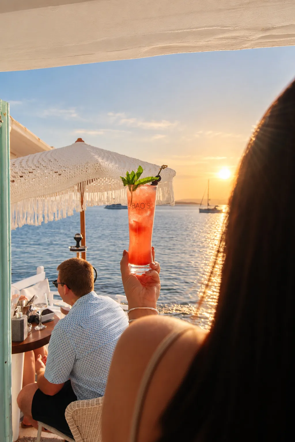 A cocktail raised against the sun going down over the sea at BAO'S