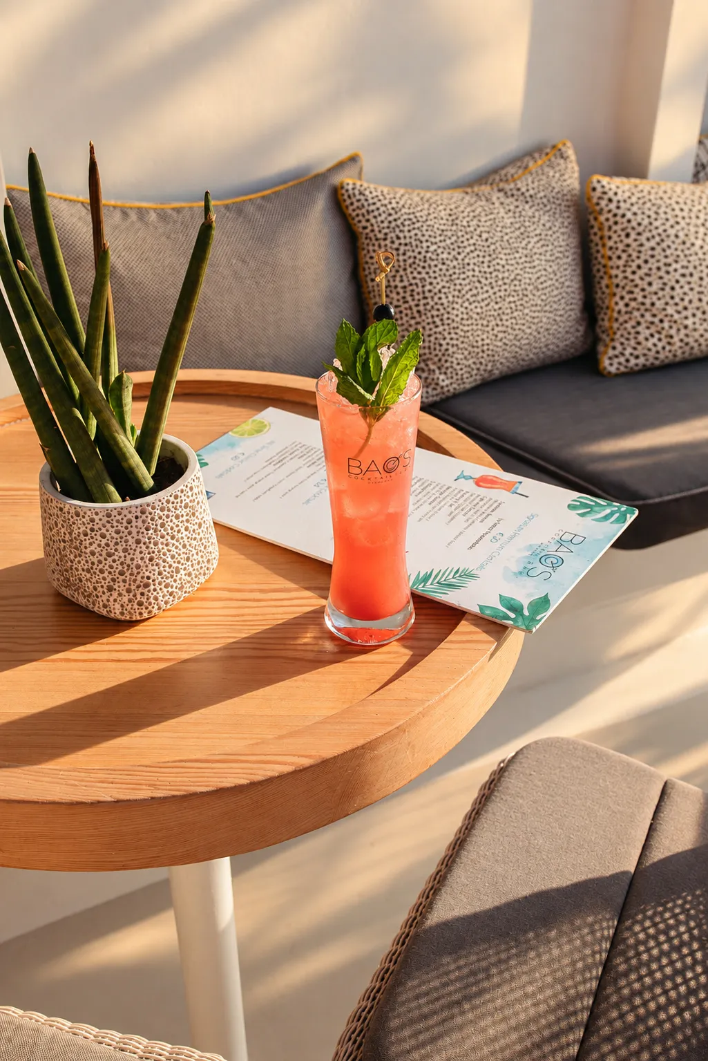 A cocktail and the menu on a wooden table at BAO'S, cushions on the bench behind and the evening light across the top