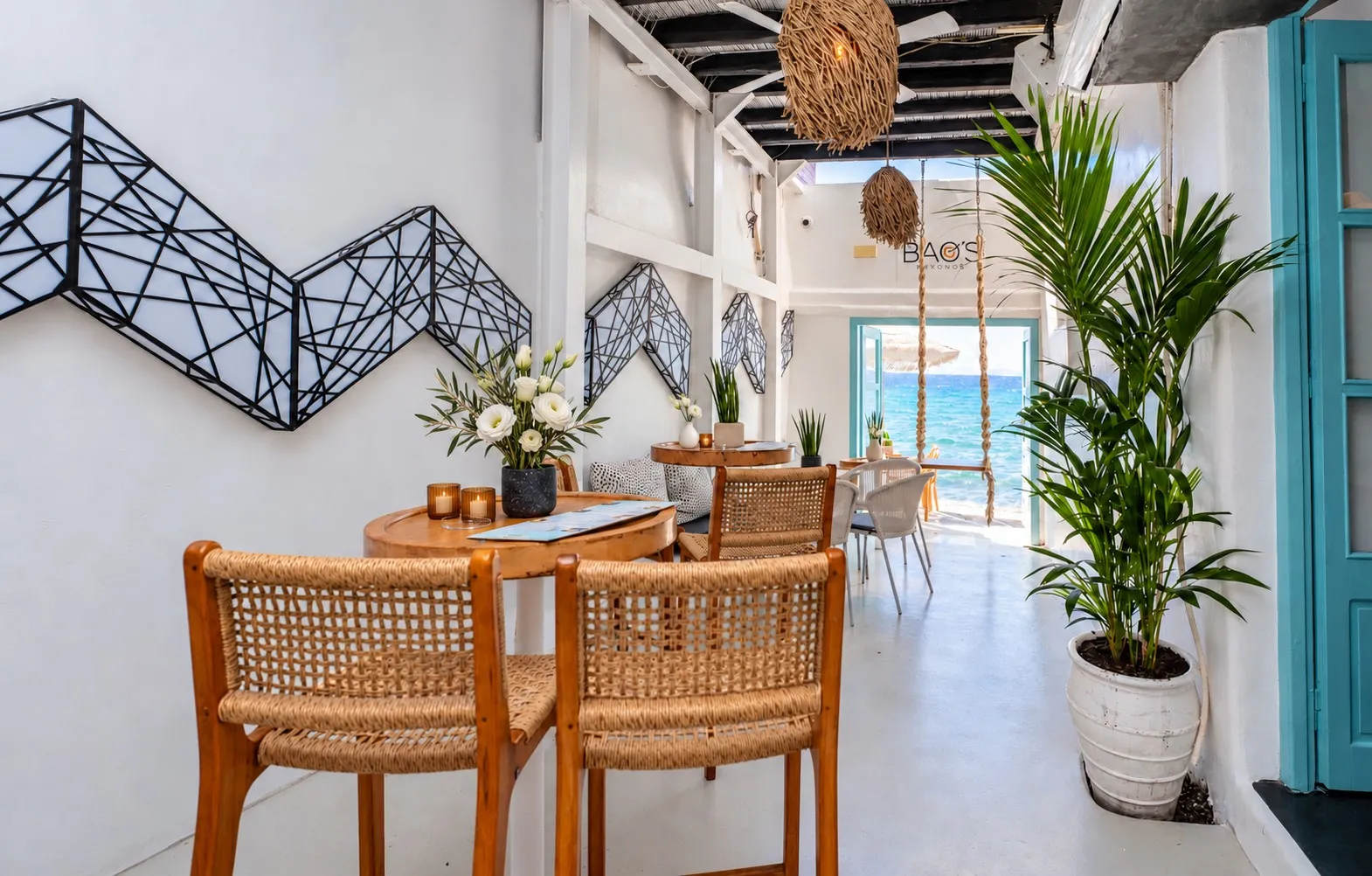 Rattan chairs at a table in the bright white lounge at BAO'S, the sea through the open door at the end