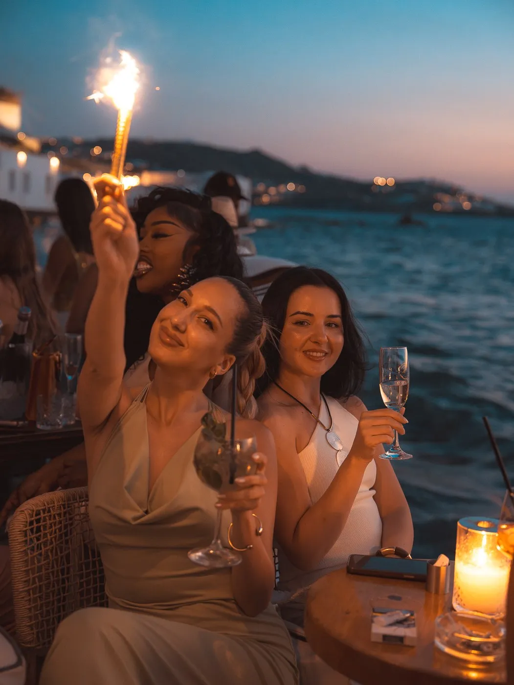 Guests at a seaside table at BAO'S holding a sparkler at dusk, the lights of the town across the water