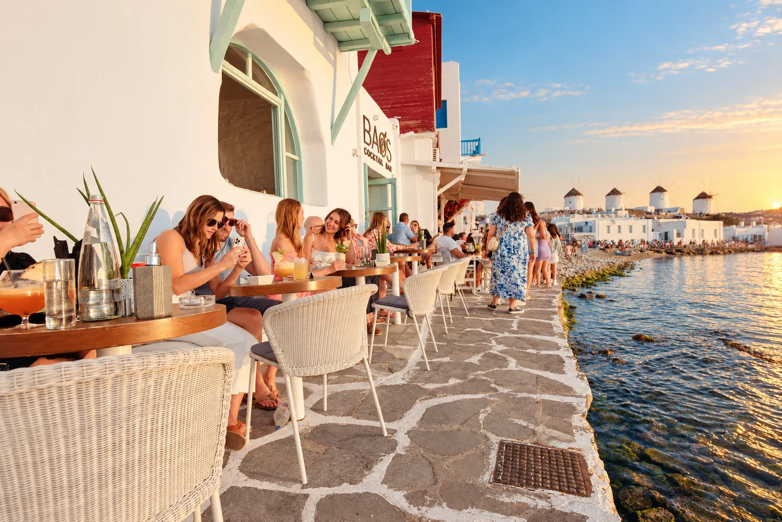 Guests at the waterfront tables at BAO'S in Little Venice at sunset, the bar's own sign above them and the windmills across the bay