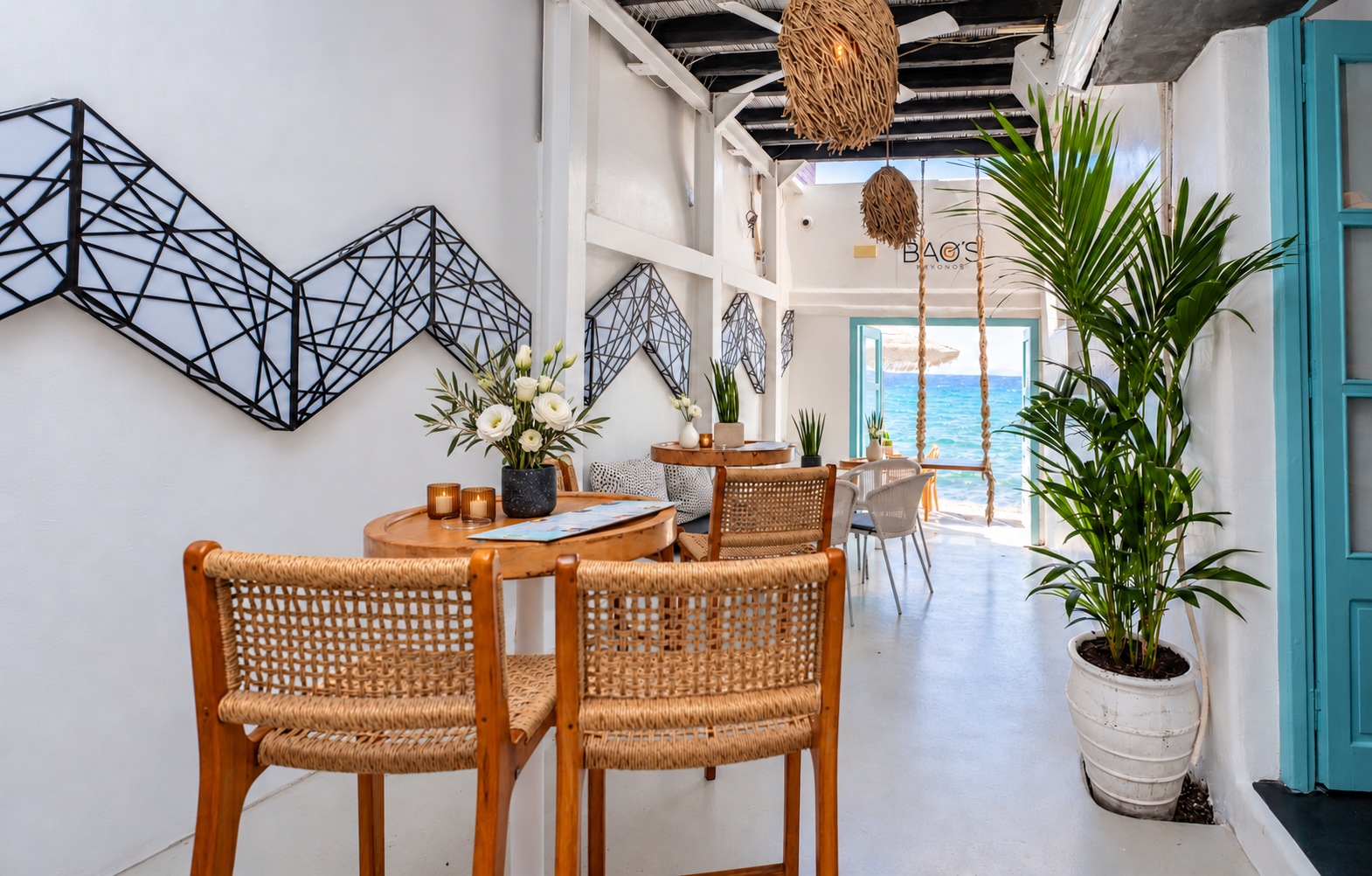 The walkway inside BAO'S in daylight, rattan chairs at a round table, the BAO'S sign and the hanging swing seat beyond, and the sea in the open doorway at the end